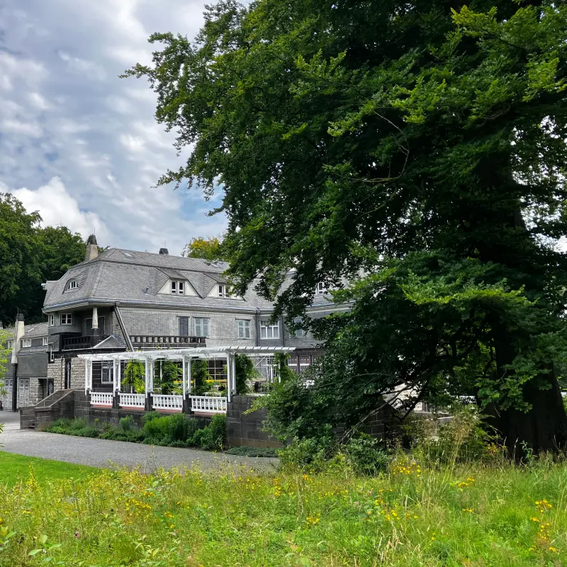 Auf dem Bild ist die Au&szlig;enansicht einer eindrucksvollen Villa inmitten einer Gartenanlage zu sehen.