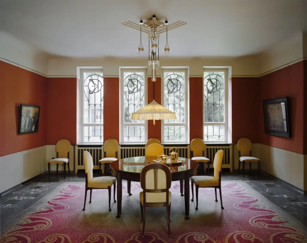 Foto: Das Speisezimmer der Villa Hohenhof mit einem Tisch und mehreren St&uuml;hlen. An den W&auml;nden h&auml;ngen zwei Gem&auml;lde und im Hintergrund sind gro&szlig;e Fenster.