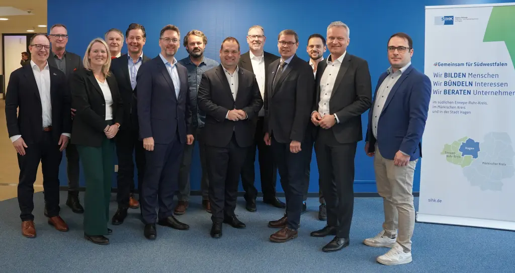 Foto: Mehrere Personen in Anz&uuml;gen stehen zusammen vor einer Wand. Daneben steht ein Roll-Up der SIHK.