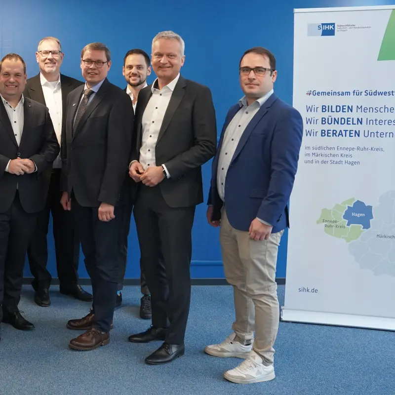 Foto: Mehrere Personen in Anz&uuml;gen stehen zusammen vor einer Wand. Daneben steht ein Roll-Up der SIHK.
