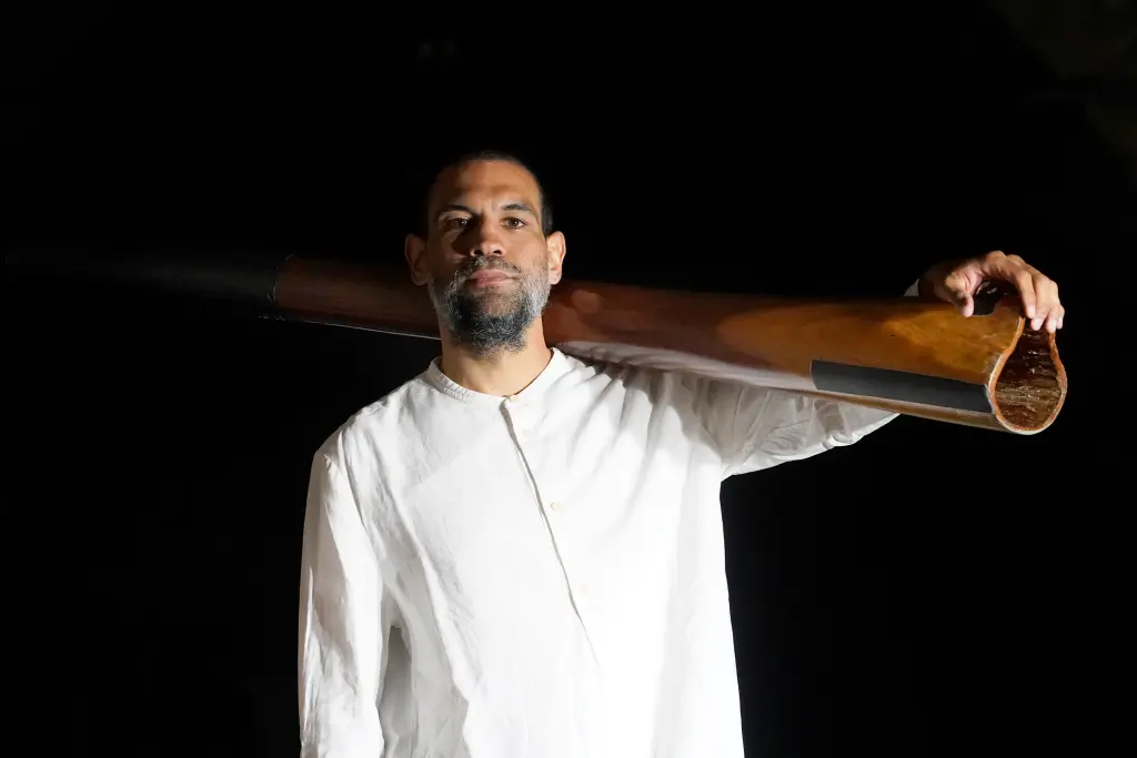 Foto: Zu sehen ist ein Mann in wei&szlig;em Gewand, der ein Didgeridoo &uuml;ber der Schulter tr&auml;gt.