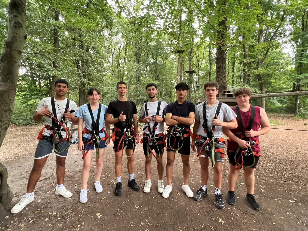 Foto: Sieben Jugendliche stehen nebeneinander in Kletterausrüstung in einem Wald und lächeln in die Kamera.
