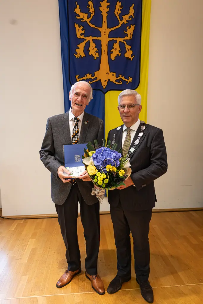 Foto: Zwei Männer mit einem Blumenstrauß und einer Urkunde in den Händen stehen im Anzug vor einer Fahne, die eine Eiche zeigt.
