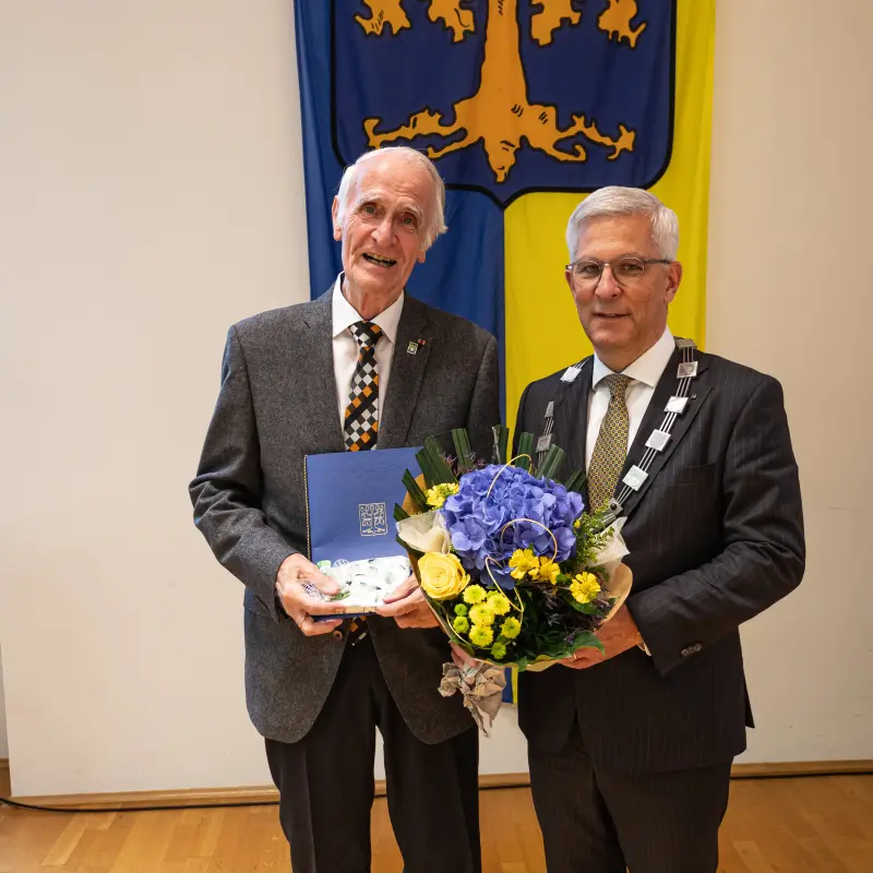 Foto: Zwei Männer mit einem Blumenstrauß und einer Urkunde in den Händen stehen im Anzug vor einer Fahne, die eine Eiche zeigt.