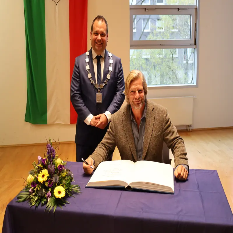 Foto: Auf einem Tisch liegt ein großes Buch. Henning Baum hat einen Stift in der Hand und schaut in die Kamera. Oberbürgermeister Dennis Rehbein trägt die Amtskette und steht neben Henning Baum.