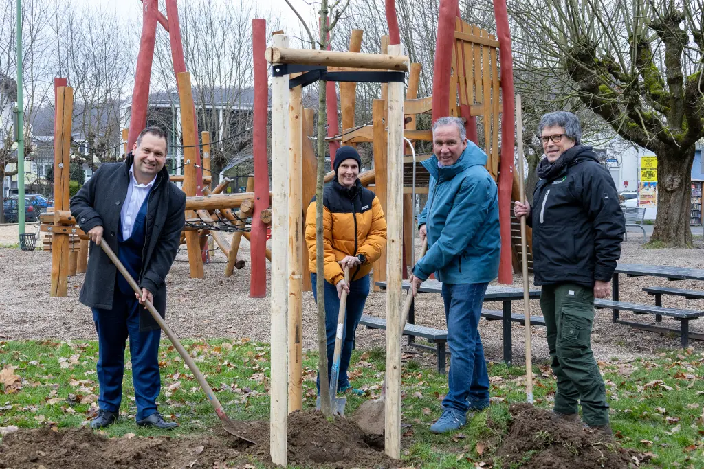Ein Gruppe von vier Menschen pflanzt gemeinsam einen Baum, alle halten Gartenwerkzeuge in den Händen. Im Hintergrund ist ein Spielplatz zu sehen.