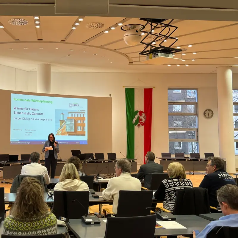 Foto: Menschen sitzen in einem gro&szlig;en Raum. Vorne steht eine Frau vor einer gro&szlig;en Leinwand.