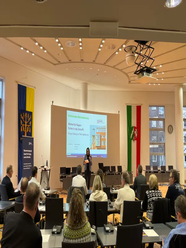 Foto: Menschen sitzen in einem gro&szlig;en Raum. Vorne steht eine Frau vor einer gro&szlig;en Leinwand.