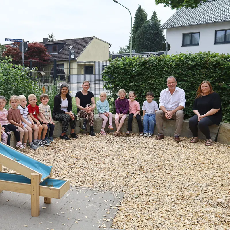 Foto: Neben dem Wasserspiel sitzen die Kinder mit den oben aufgez&auml;hlten Personen.
