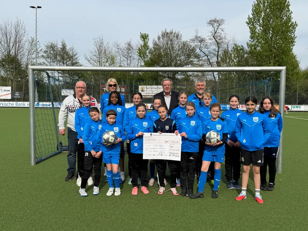 Foto: Eine M&auml;dchengruppe und vier Erwachsene stehen mit einem Scheck auf einem Fu&szlig;ballplatz vor einem Tor.
