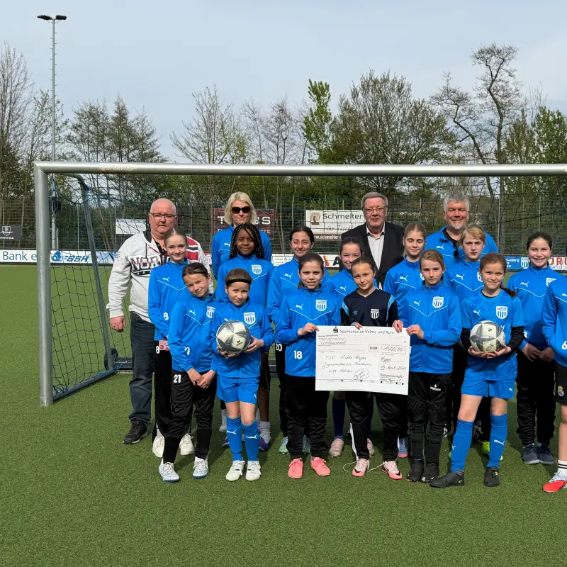 Foto: Eine M&auml;dchengruppe und vier Erwachsene stehen mit einem Scheck auf einem Fu&szlig;ballplatz vor einem Tor.