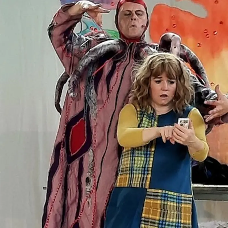 Foto: Eine Frau und ein Mann f&uuml;hren in bunten Kost&uuml;men etwas auf. Die Frau hat ein Handy in der Hand und der Mann h&auml;lt seine H&auml;nde bedrohlich vor und hinter sie.