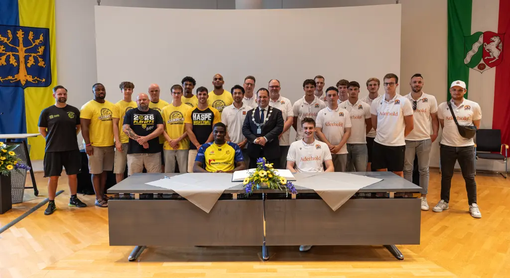 Foto: Ein Gruppenfoto mit M&auml;nnern in Trikots mit zwei Tischen im Vordergrund in einem Saal. Ein Mann tr&auml;gt einen Anzug und eine Amtskette.