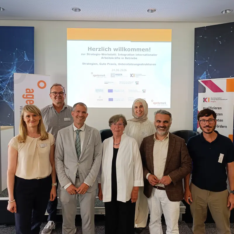 Foto: Die onem aufgef&uuml;hrten Personen stehen aufgereiht neben- und hintereinander, im Hintergrund ist eien Pr&auml;&auml;sentation zu sehen. Neben ihnen stehen Roll-Up-Banner.