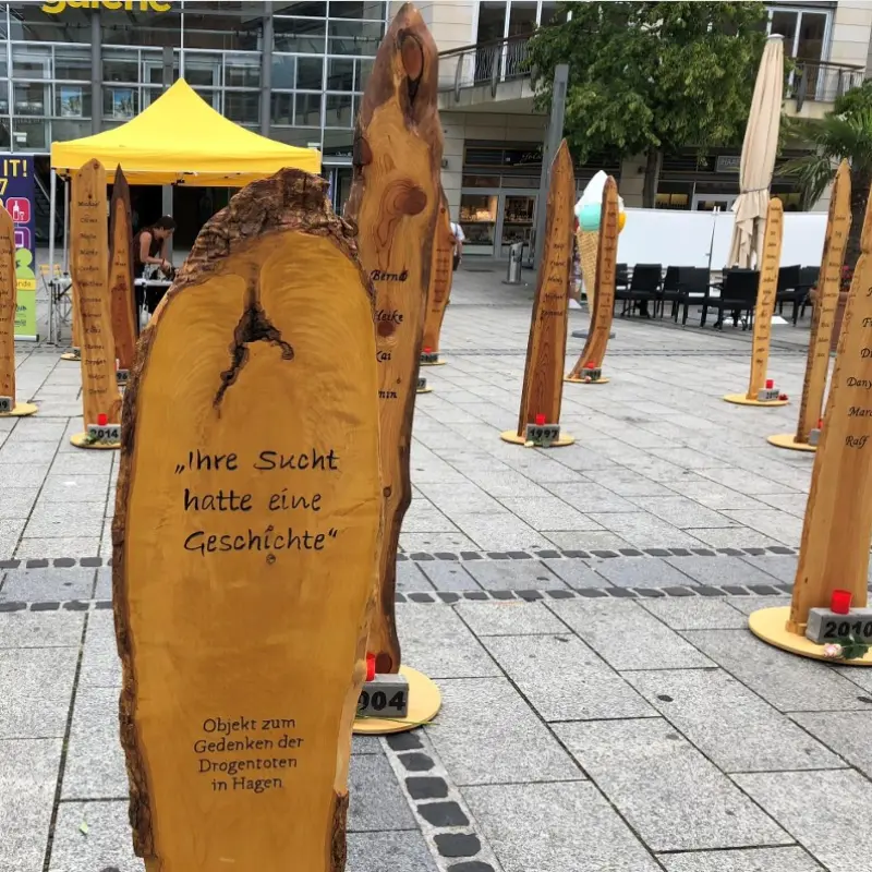 Foto: Holzstelen, auf denen Namen notiert wurden. Darunter steht in Stein eingemei&szlig;elt jeweils ein Jahr. Auf einer Stele setht "Ihre Sucht hatte eine Geschichte" - Objekt zum Gedenken der Drogentoten in Hagen.