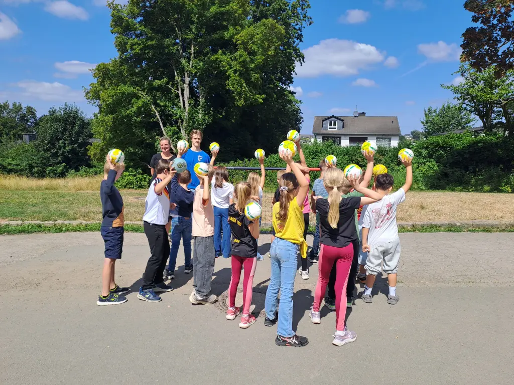 Foto: Die oben genannten Personen stehen mit Kindern, die Handbälle in der Hand halten, vor einem Tor.