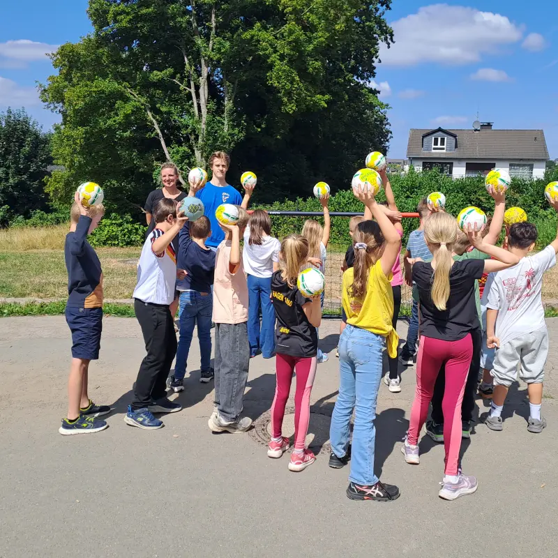 Foto: Die oben genannten Personen stehen mit Kindern, die Handbälle in der Hand halten, vor einem Tor.