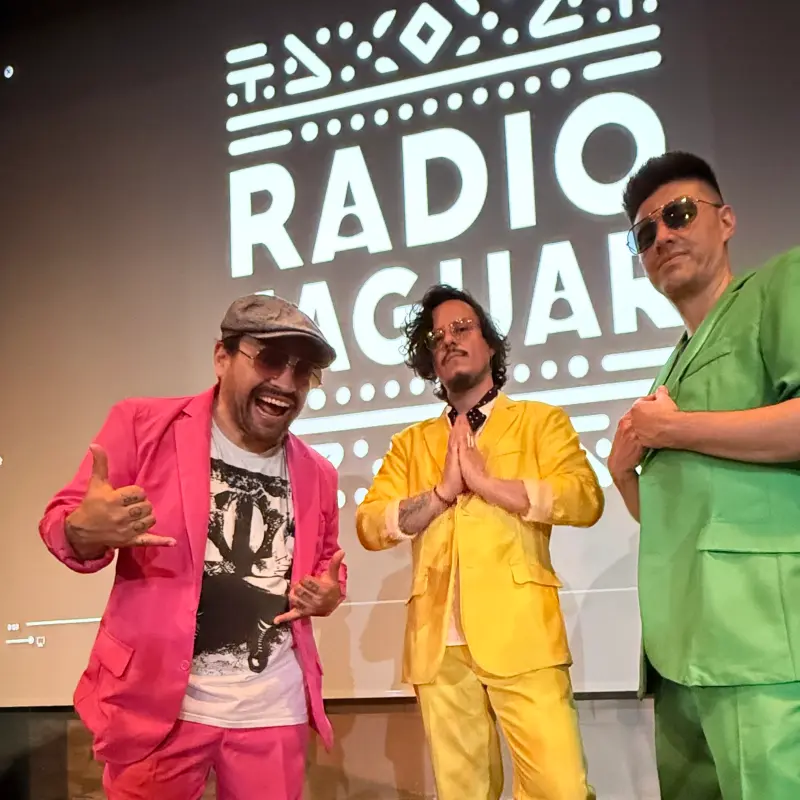 Foto: Die Bandmitglieder haben knallbunte Anzüge an und stehen vor einer Wand, auf der "Radio Jaguar" steht.