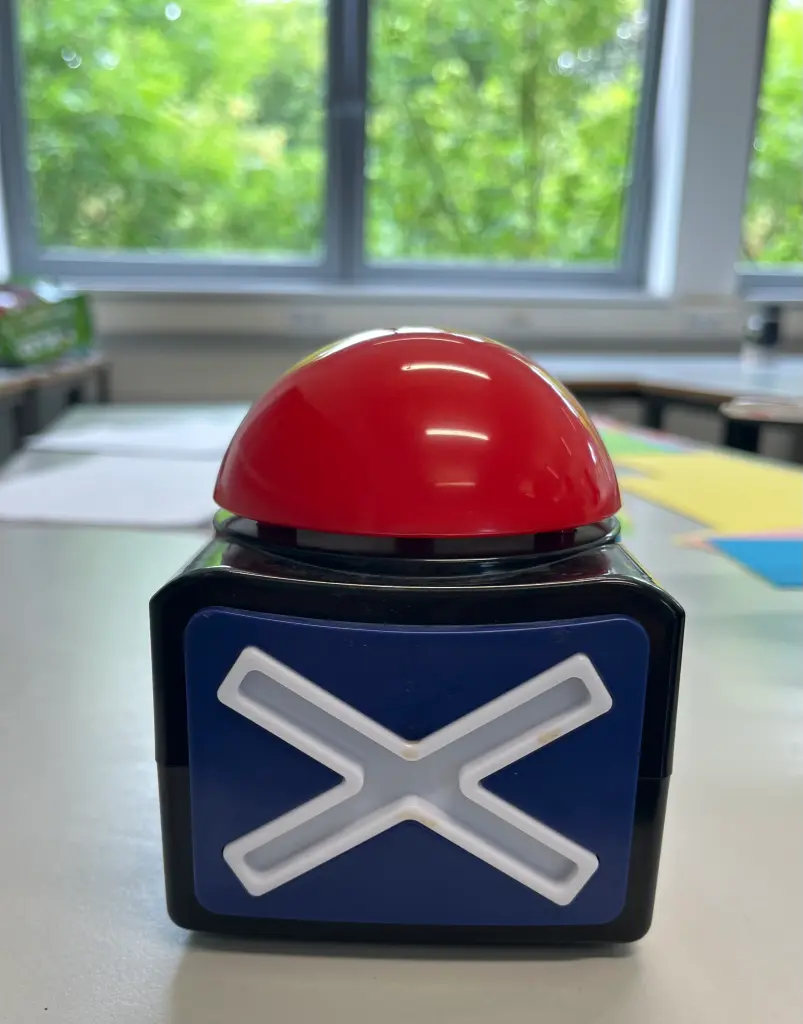 Foto: Ein roter Buzzer mit einem Kreuz an der Seite steht auf einem Tisch.