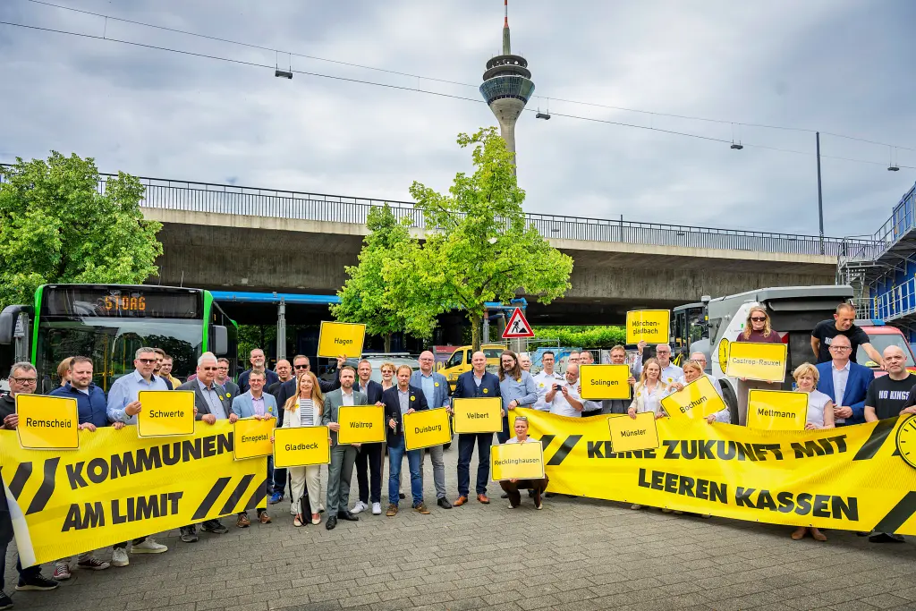 Foto: Vertreter der Kommunen stehen vor einem Bus, einer Kehrmaschine, einem Feuerwehrauto und halten Schilder mit Ortsnamen hoch sowie Banner mit der Aufschrift "Kommunen am Limit" sowie "Keine Zukunft mit leeren Kassen"