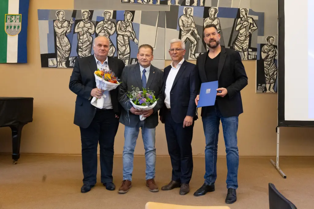 Foto: Vier Personen stehen nebeneiander zur Kamera gerichte, zwei halte Blumensträuße, einer ein blaues Buch.