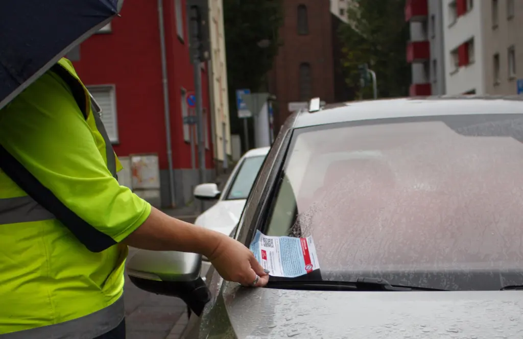 Foto: Eine Ordnungsbeamtin mit gelber Weste klemmt einen Strafzettel an ein Auto.