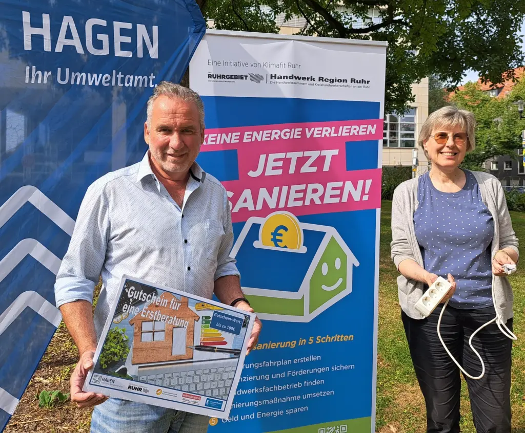 Foto: Ein Mann und eine Frau stehen vor einem Rollamt mit der Aufschrift "Keine Energie verlieren - Jetzt sanieren!". Der Mann hält einen großen beispielhaften Gutschein für eine Erstberatung in den Händen.