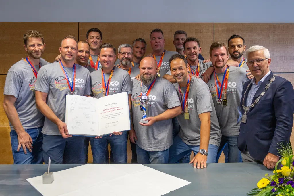 Foto: Zu sehen sind mehrere Menschen mit grauen Shirts und Medaillen um den Hals, zwei davon halten ein aufgeschlagenes Buch mit Unterschriften, einer einen Pokal. Rechts befindet sich Oberbürgermeister Erik O. Schulz im Anzug.