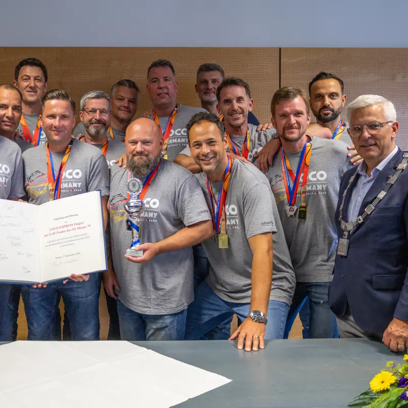 Foto: Zu sehen sind mehrere Menschen mit grauen Shirts und Medaillen um den Hals, zwei davon halten ein aufgeschlagenes Buch mit Unterschriften, einer einen Pokal. Rechts befindet sich Oberbürgermeister Erik O. Schulz im Anzug.