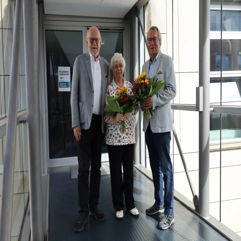 Foto: Drei Personen, zwei davon mit Blumensträußen stehen nebeneinander und lächeln in die Kamera.