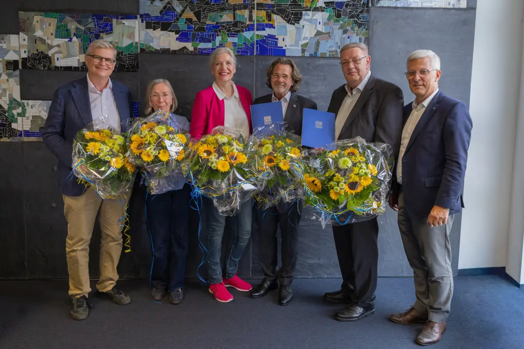 Foto: Drei Männer und drei Frauen in schicker Kleidung lächeln in die Kamera. Bis auf einen halten alle Personen einen Blumenstrauß in der Hand.