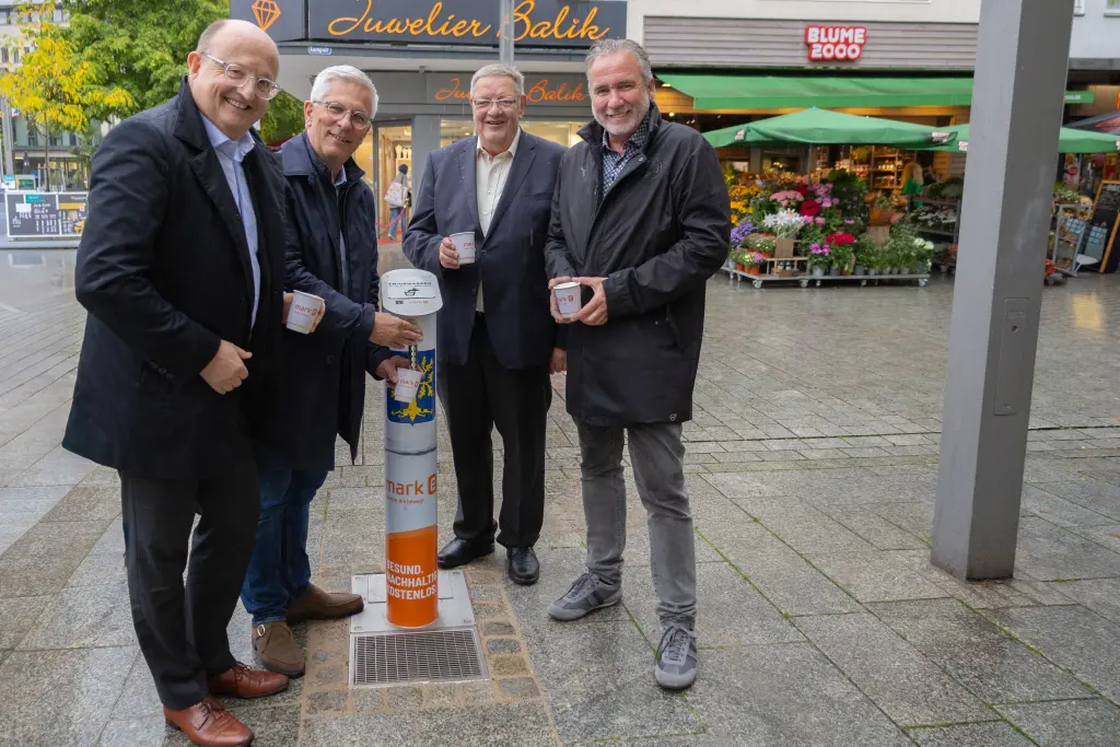 Foto: Vier Männer stehen neben einem kleinen Trinkwasserbrunnen in der Hagener Innenstadt.