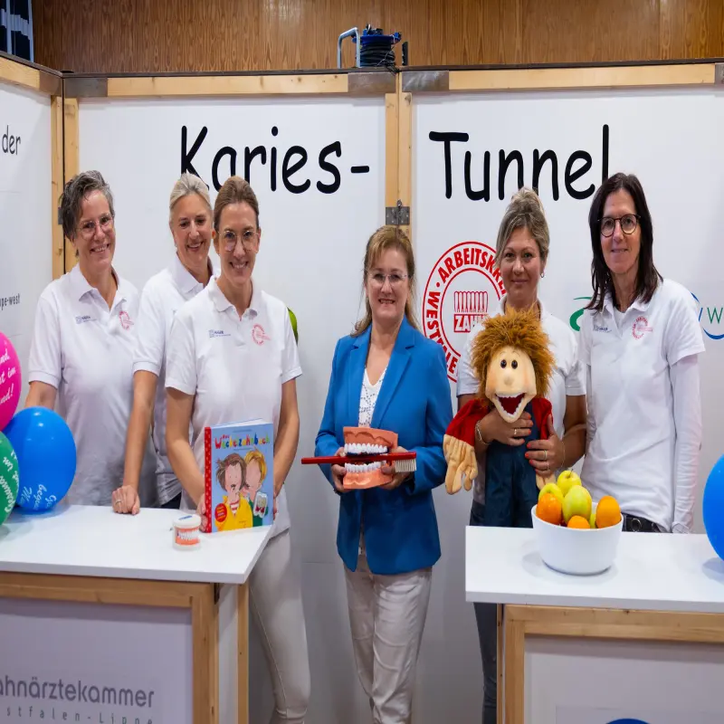 Foto: Sechs Frauen, fünf davon mit weißen T-Shirts, lächeln in die Kamera. Die Frau in der Mitte hält ein großes Plastik-Gebiss und eine große Zahnbürste in den Händen