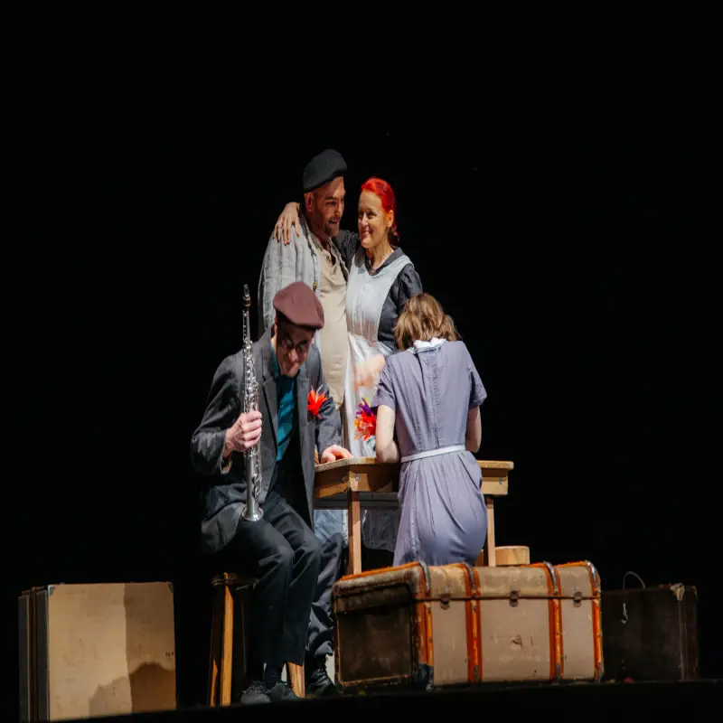 Foto: Zu sehen ist eine Szene eines Theaterstücks, bei der sich vier Personen in bäuerlicher Kleidung an einem Tisch aufhalten. Zwei unterhalten sich, ein anderer hält ein Blasinstrument in der Hand.