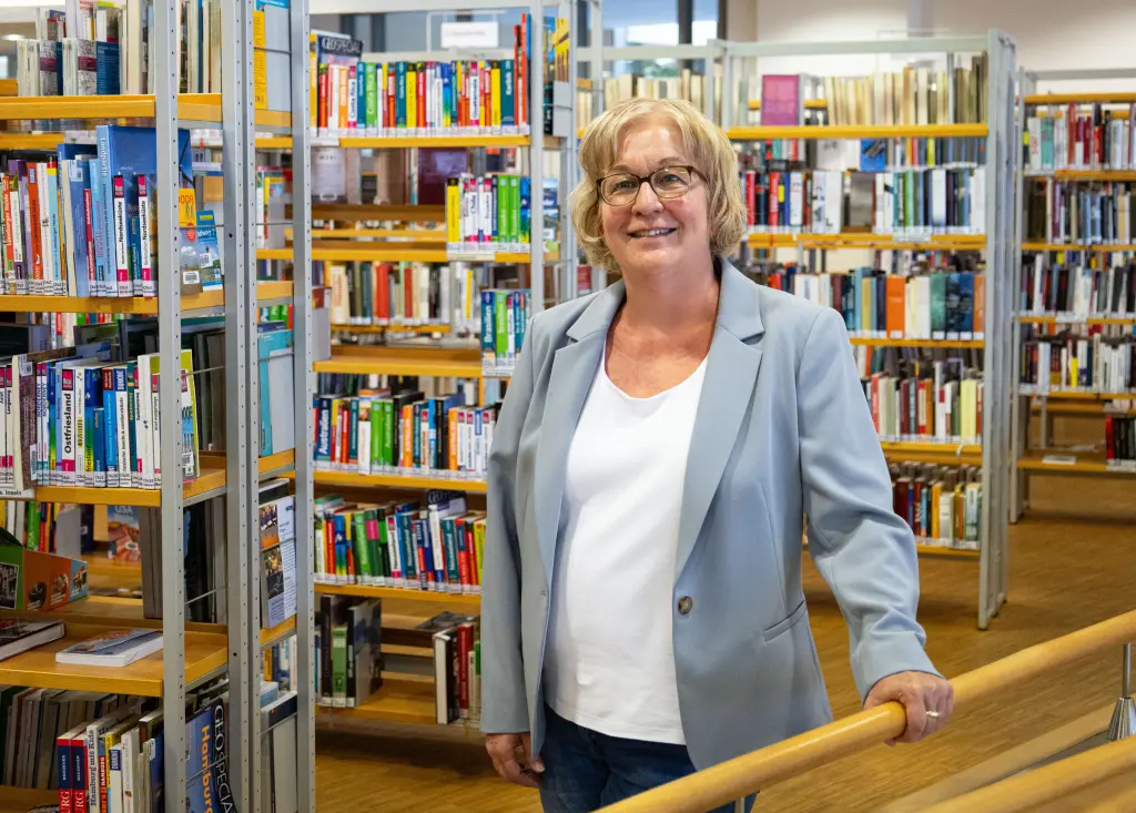 Foto: Eine Dame lächelt freundlich in die Kamera, im Hintergrund befinden sich Bücherregale.