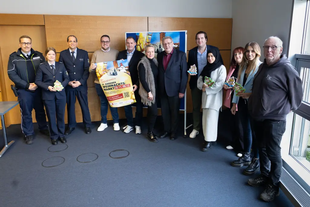 Foto: Viele Personen stehen nebeneinander und lächeln in die Kamera. Sie präsentieren das Weihnachtsmarktplakat sowie mehrere Lebkuchenbäume.