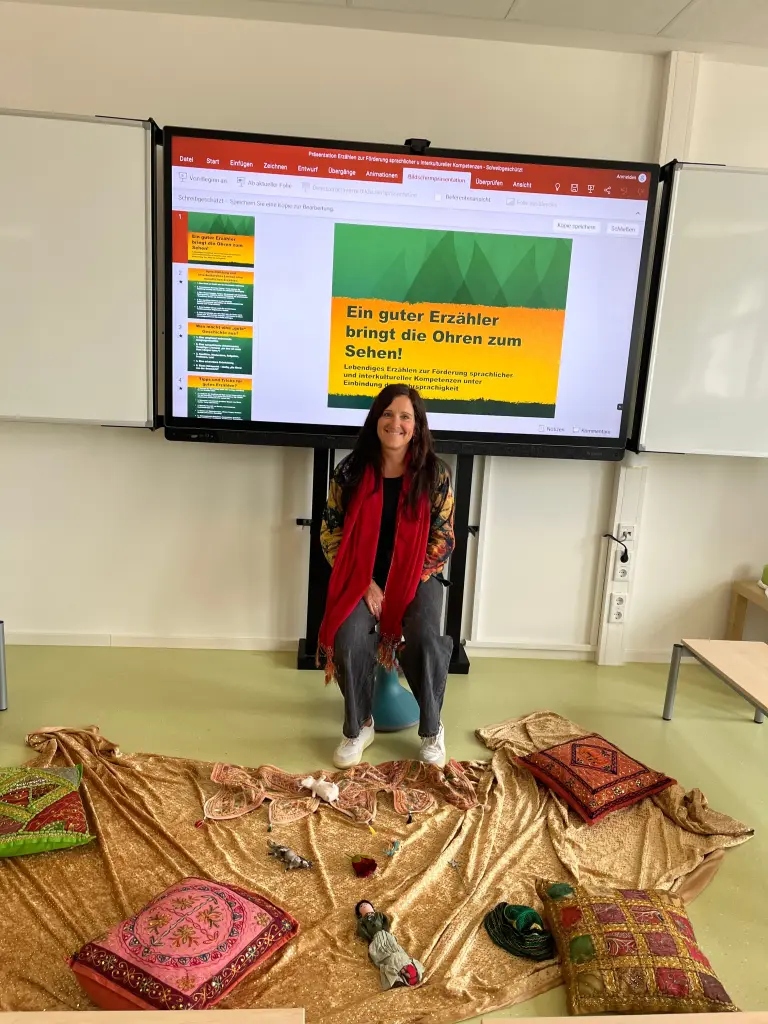 Foto: Workshop-Leiterin Katrin Petri sitzt vor einer Powerpoint-Präsentation über das Thema. Vor ihr liegt eine Decke mit Kissen und kleinen Gegenständen, zum Beispiel eine Puppe.