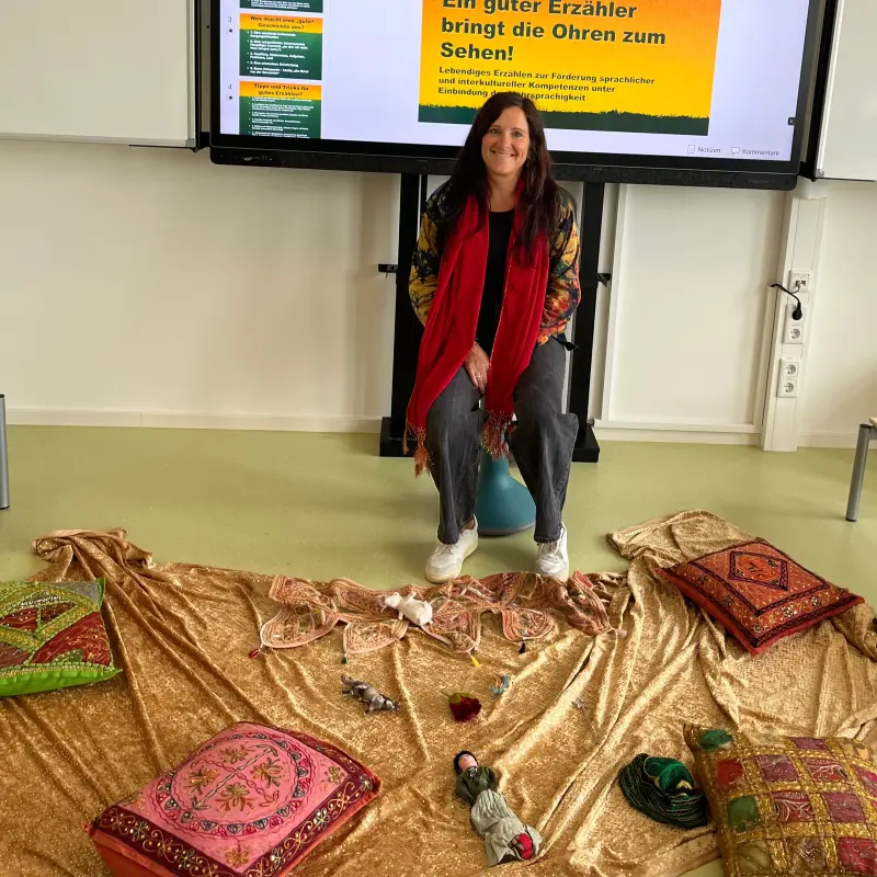 Foto: Workshop-Leiterin Katrin Petri sitzt vor einer Powerpoint-Präsentation über das Thema. Vor ihr liegt eine Decke mit Kissen und kleinen Gegenständen, zum Beispiel eine Puppe.