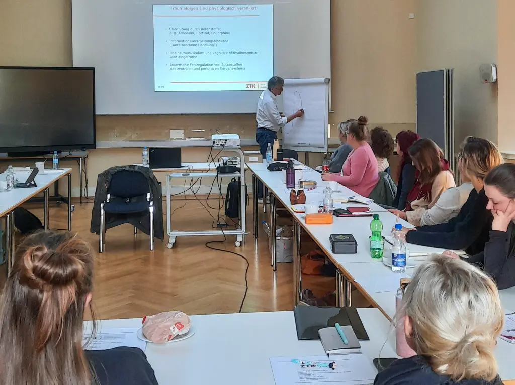 Foto: Mehrere Personen sitzen an einem Tisch in einem Seminarraum, vorne zeichnet der Dozent auf einem Flipchart.