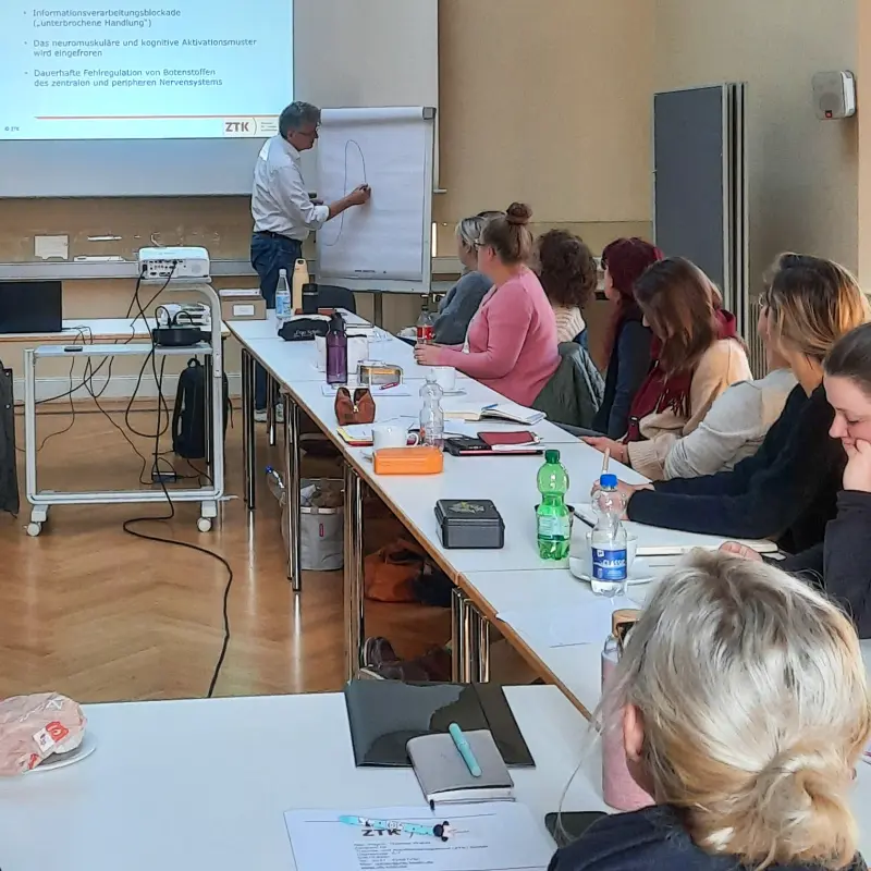 Foto: Mehrere Personen sitzen an einem Tisch in einem Seminarraum, vorne zeichnet der Dozent auf einem Flipchart.