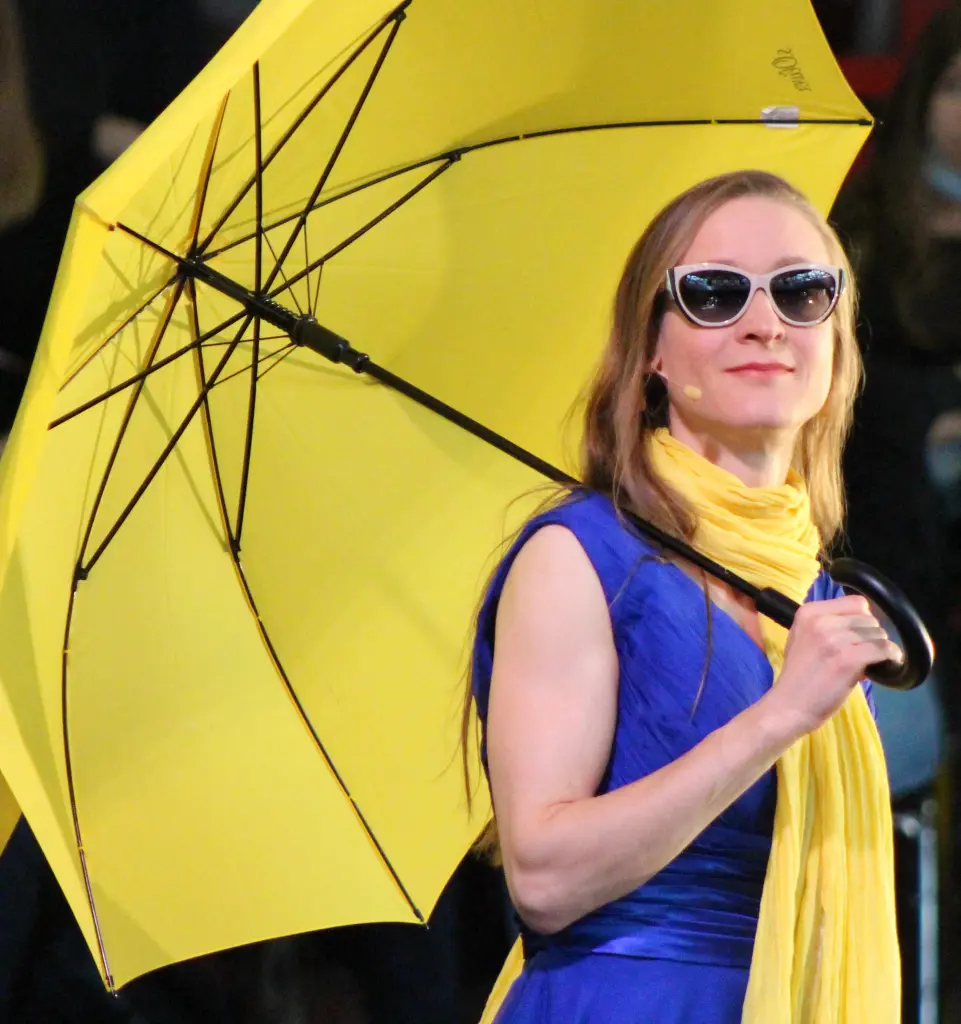 Foto: Zu sehen ist eine Frau mit blauem Kleid, gelbem Schal, gelbem Regenschirm und Sonnenbrille.
