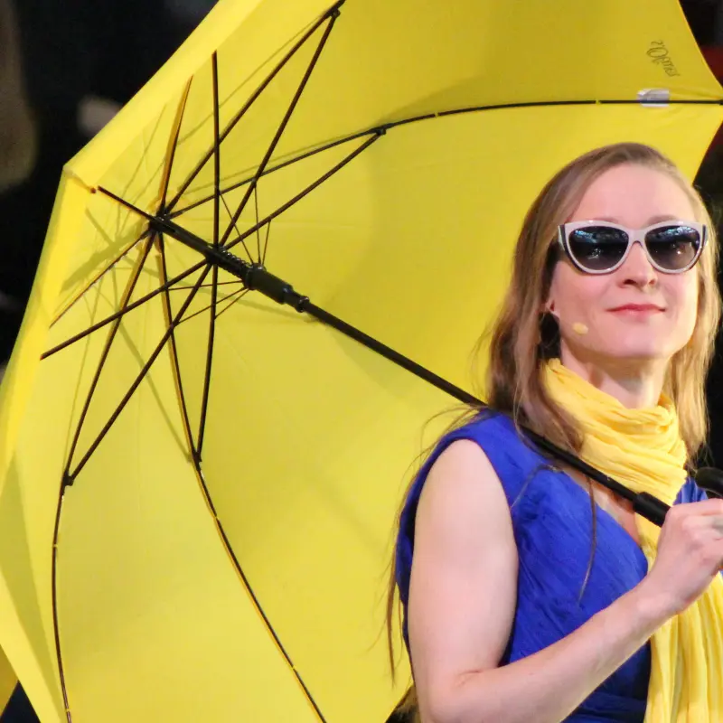 Foto: Zu sehen ist eine Frau mit blauem Kleid, gelbem Schal, gelbem Regenschirm und Sonnenbrille.