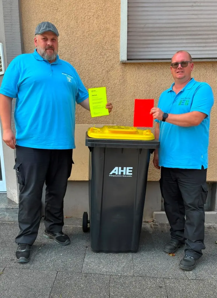Foto: Zwei Waste Watcher stehen neben einer Wertstofftonne und halten jeweils eine gelbe und eine rote Hinweiskarte in den Händen.