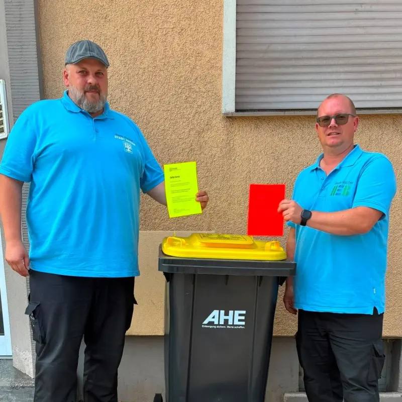 Foto: Zwei Waste Watcher stehen neben einer Wertstofftonne und halten jeweils eine gelbe und eine rote Hinweiskarte in den Händen.