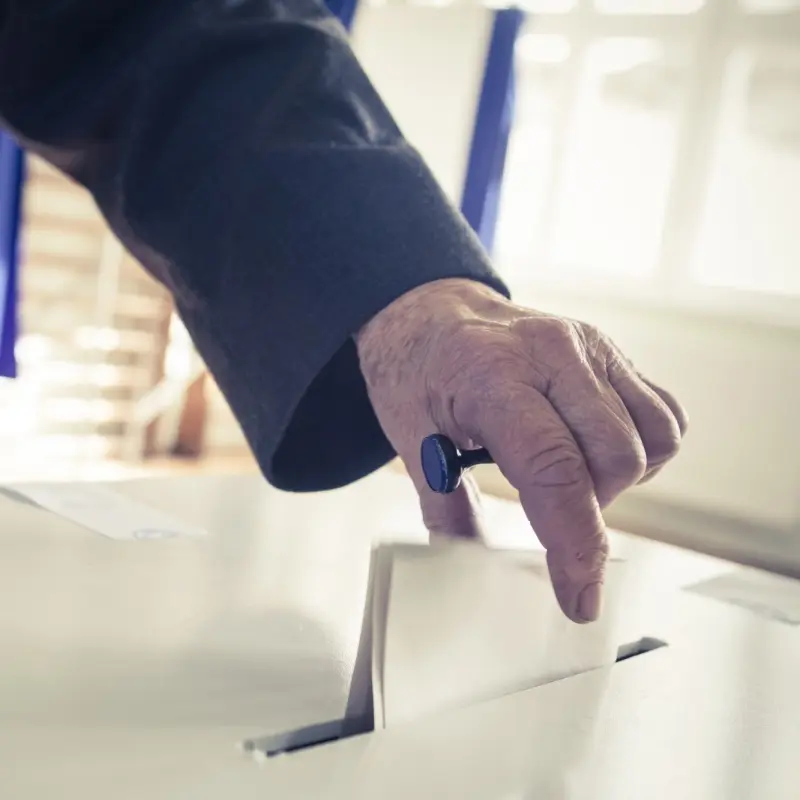 Foto: Nahaufnahme einer Hand, die einen gefalteten Zettel in eine Wahlurne steckt.