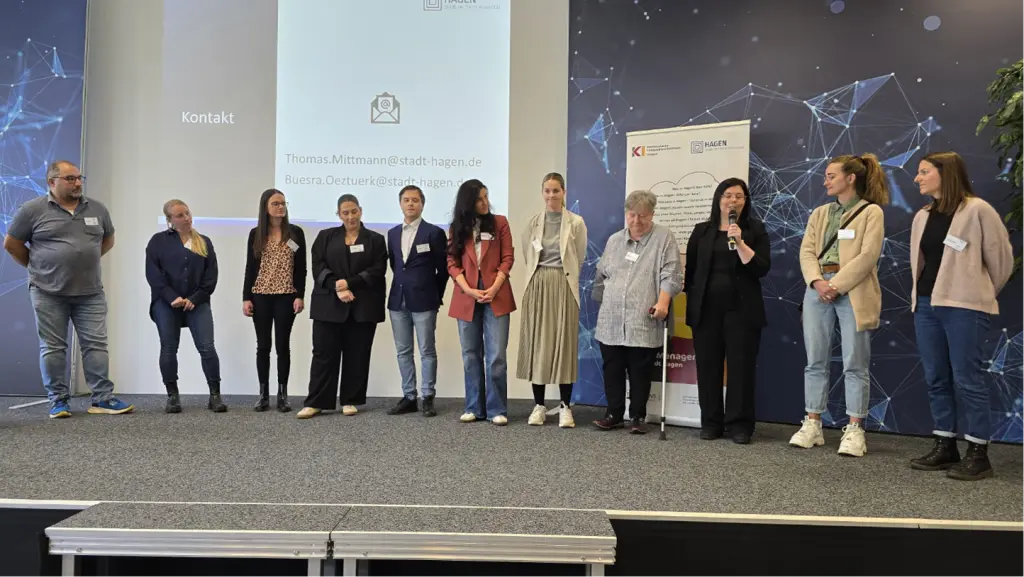 Foto: Viele Personen mit Namensschilder stehen auf einer Bühne, eine von ihnen redet in ein Mikrofon.