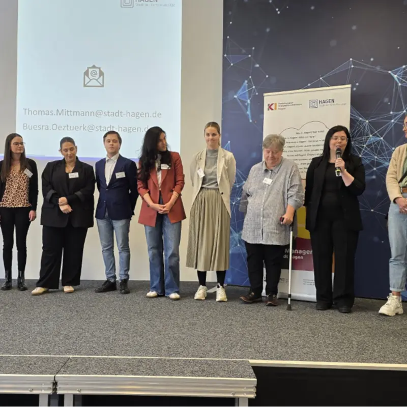 Foto: Viele Personen mit Namensschilder stehen auf einer Bühne, eine von ihnen redet in ein Mikrofon.