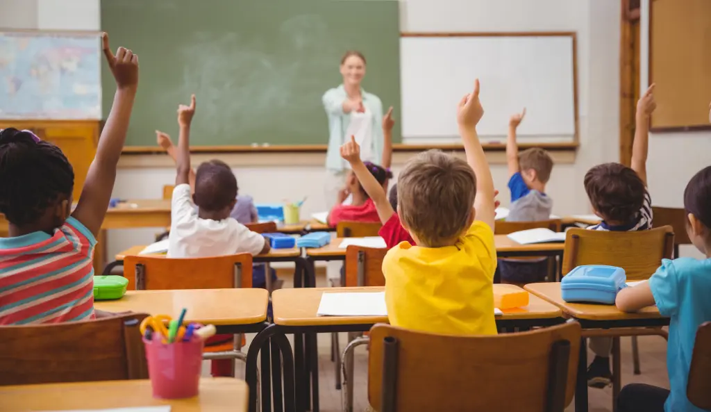 Foto: Eine Lehrerin steht in einem Klassenzimmer vor einer Tafel, im Klassenzimmer sitzen viele Kinder, die sich melden.