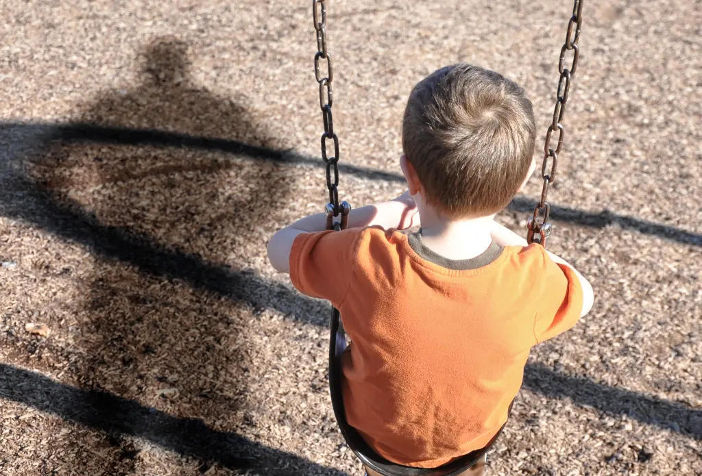 Foto: Ein Kind sitzt mit dem Rücken zur Kamera auf einer Schaukel, über ihm sieht man den Schatten einer erwachsenen Person.