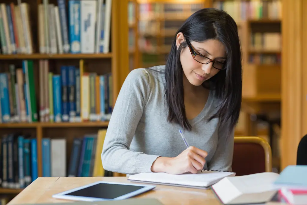 Foto: Eine Frau mit Brille sitzt an einem Tisch und macht sich Notizen, im Hintergrund stehen Bücherregale